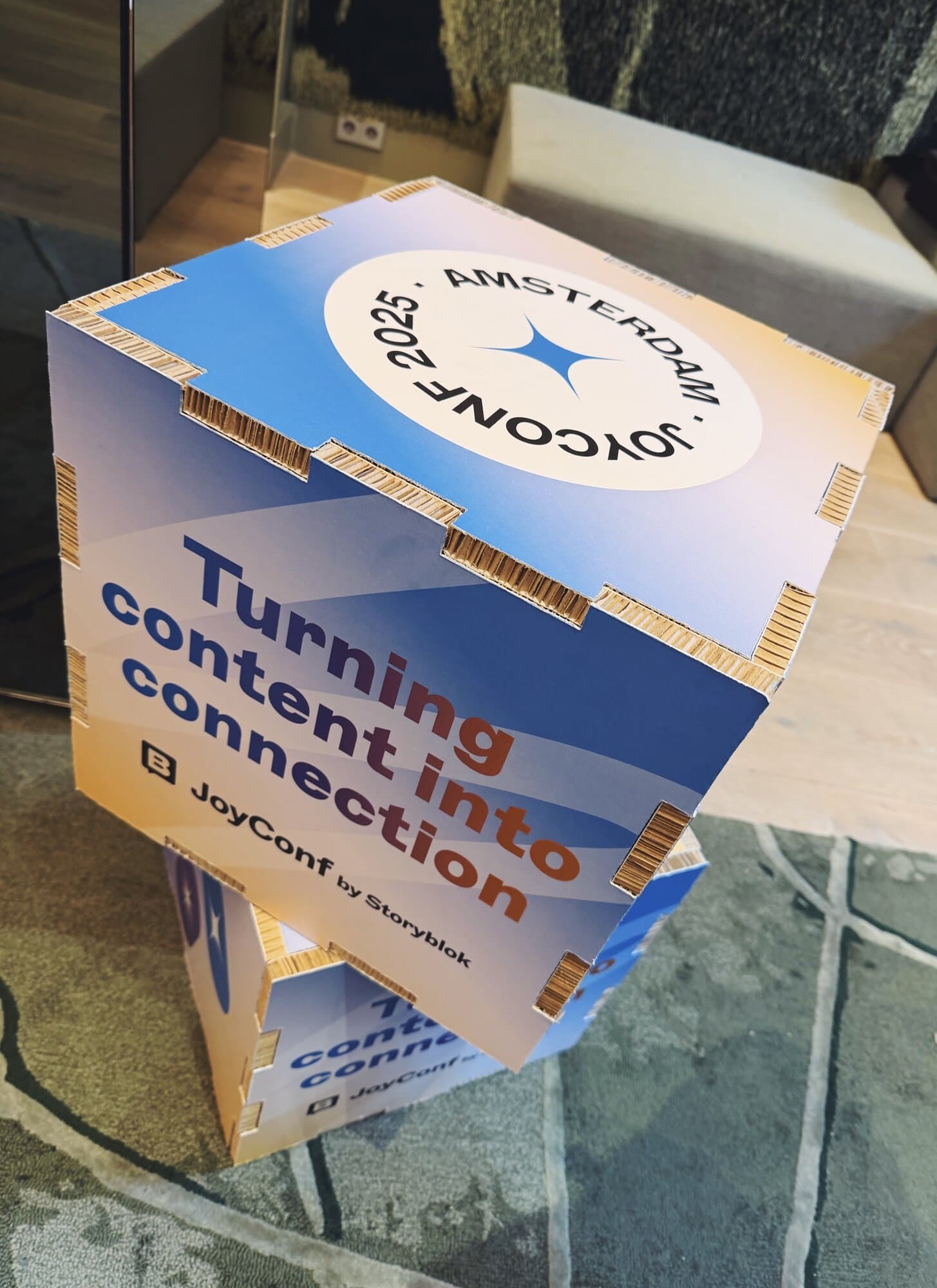 Stacked cardboard boxes with "Turning content into connection" and "JoyConf Amsterdam 2025" printed on them, on a carpeted floor.