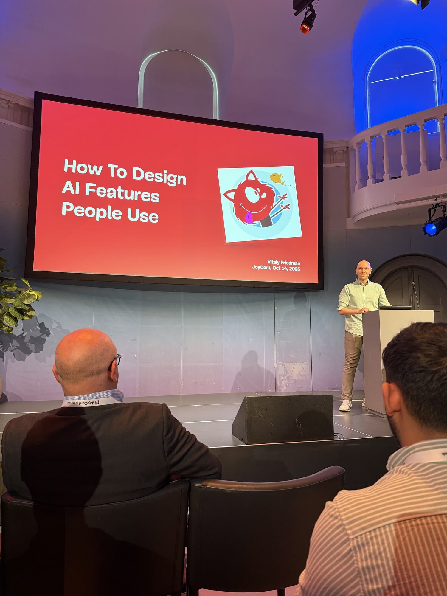 Vitaly Friedman Smashing Magazine founder presents "How To Design AI Features People Use" on a large screen at a conference venue with an audience seated.