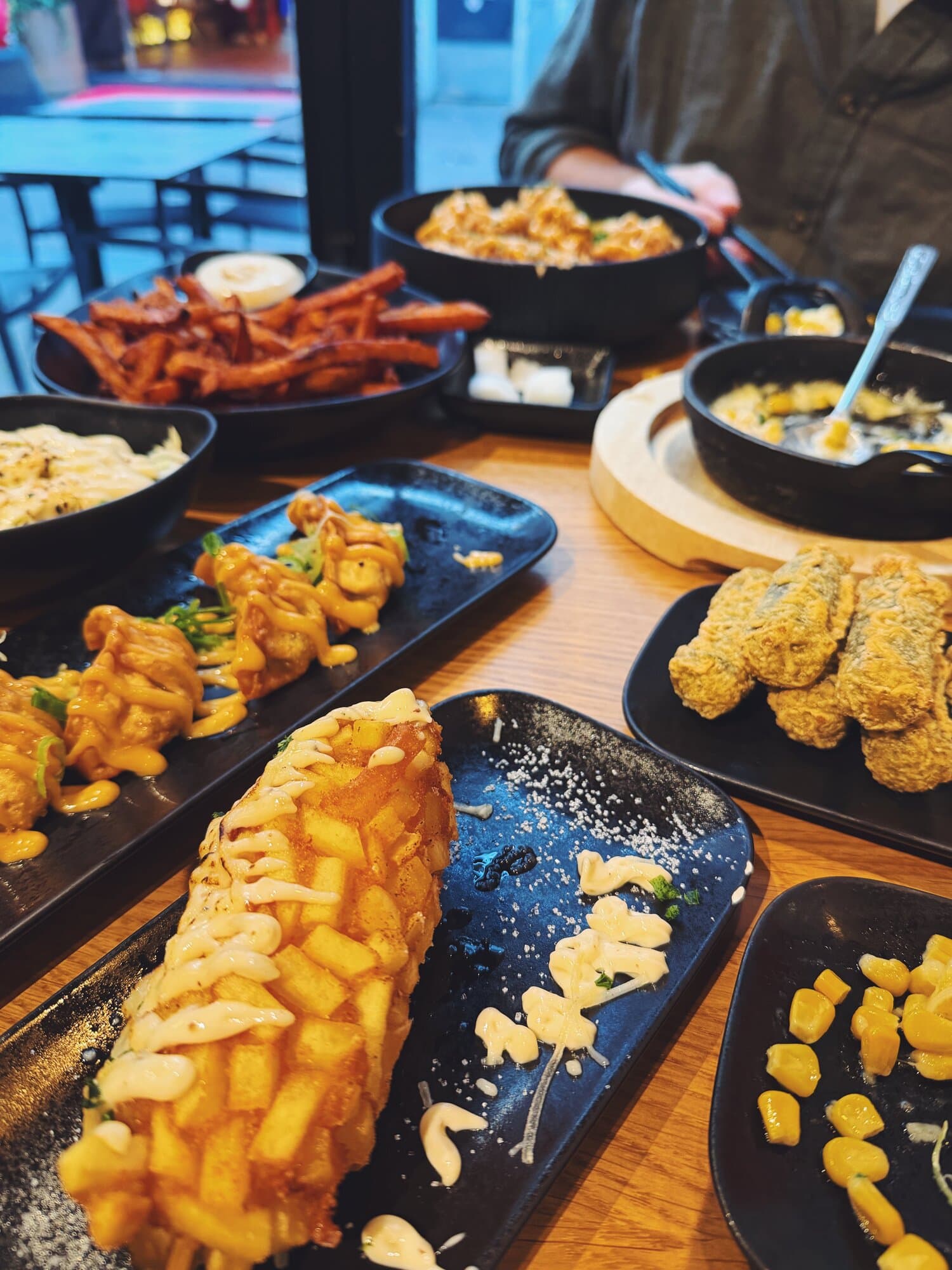 A table filled with various dishes including battered corn dogs, fried chicken, sweet potato fries, corn, and other appetizers in black plates at Hongdae Amsterdam.