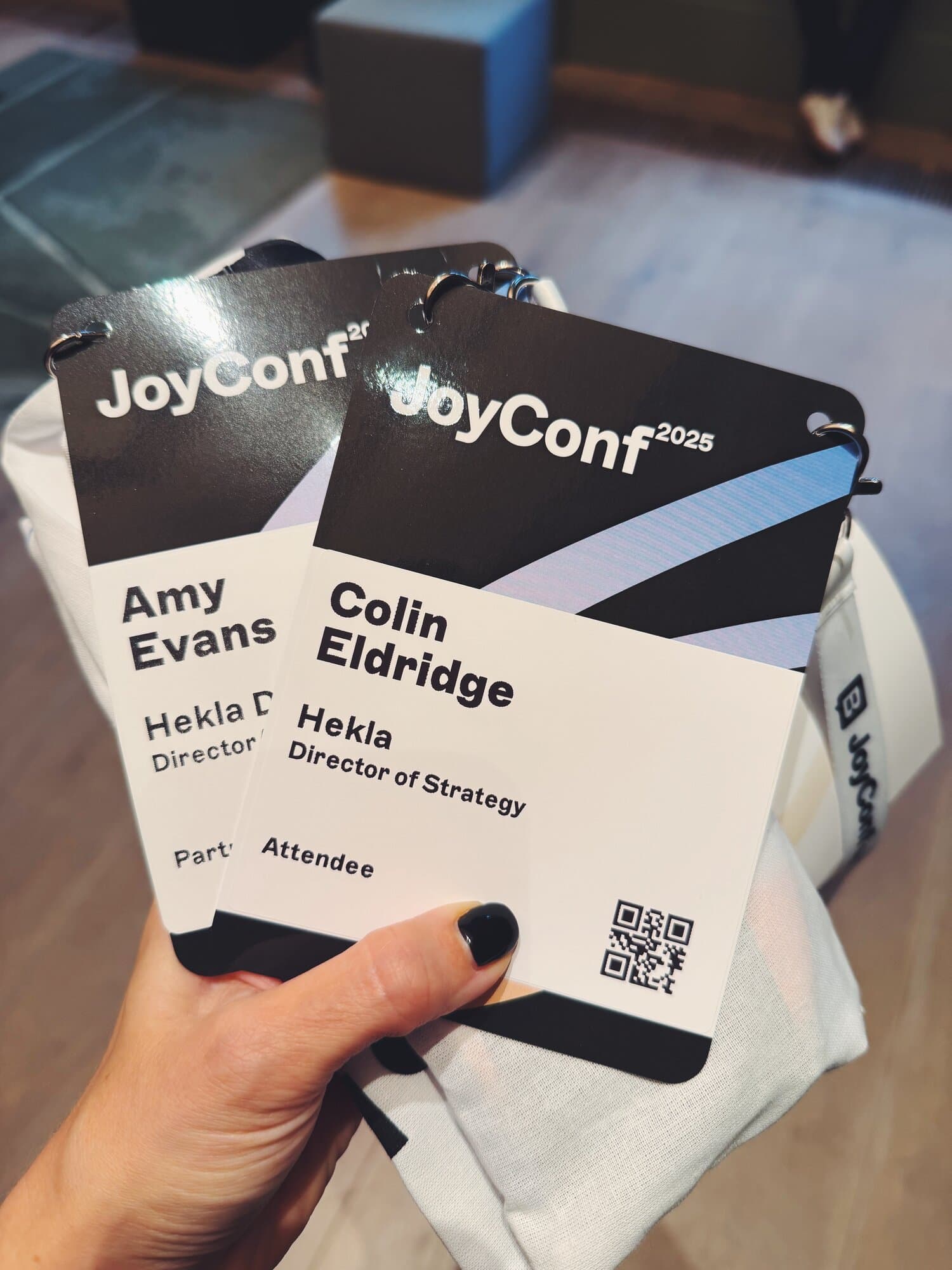 Close-up of two JoyConf 2025 attendee badges held in a hand, displaying names and titles. Background shows a blurry indoor setting.