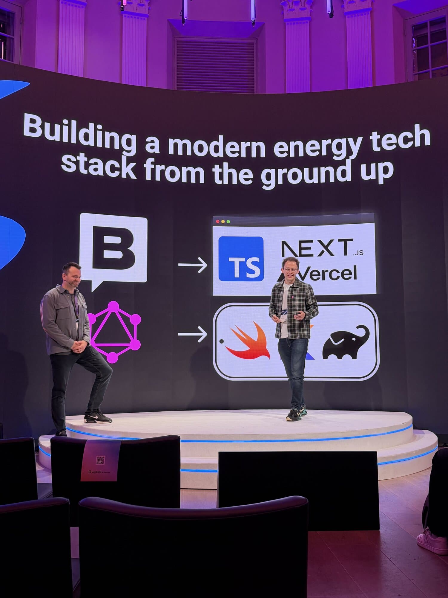 Two presenters stand on stage discussing a modern energy tech stack with visuals of programming tools on a large screen behind them.