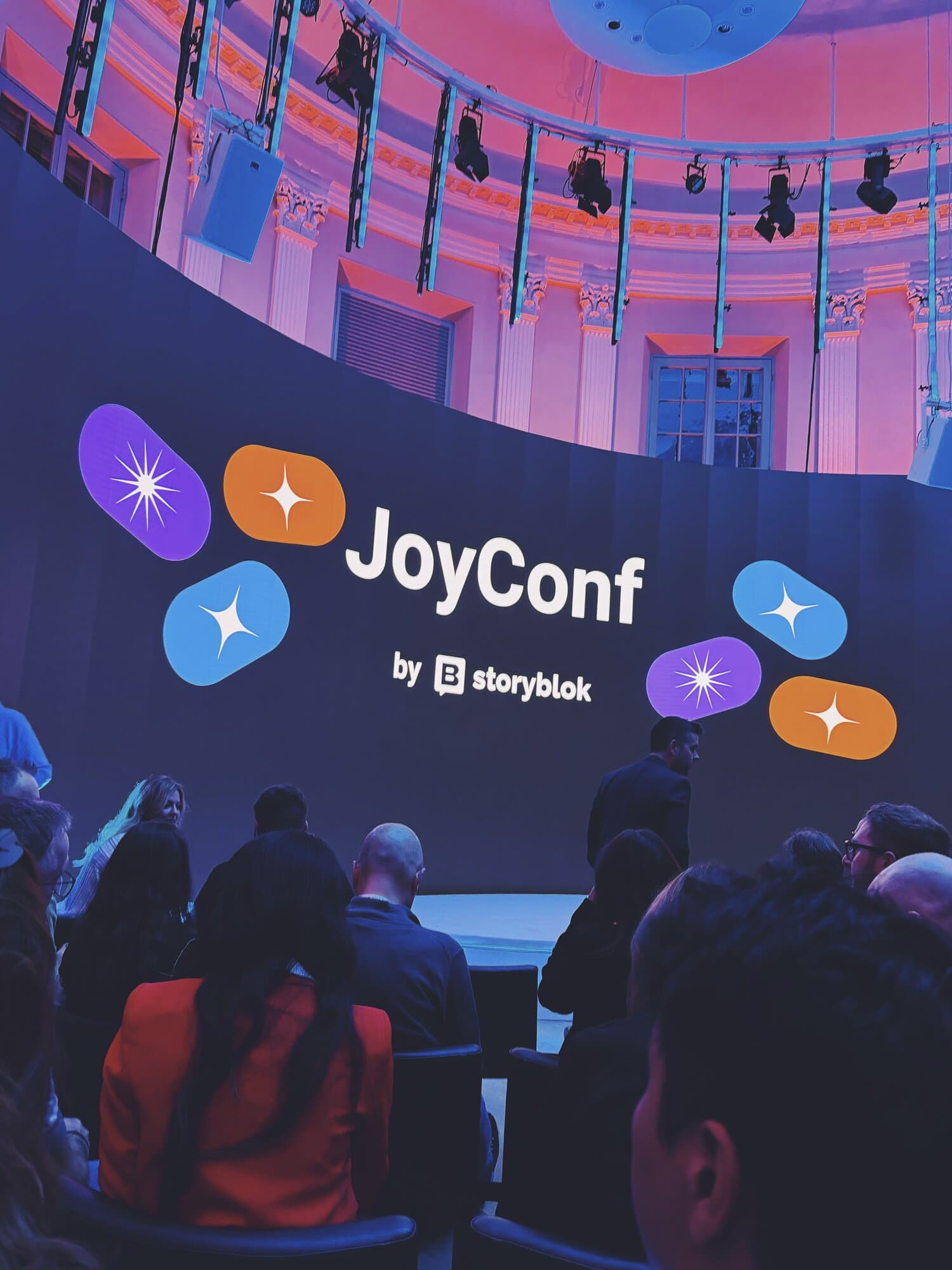 People sitting and standing at a conference with a large curved screen displaying "JoyConf by Storyblok" in a modern venue with pink lighting.
