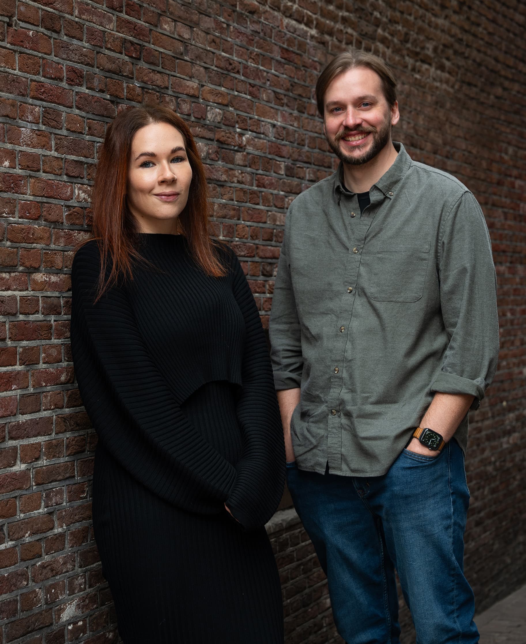 Amy and Colin. A woman in a black dress and a man in a green shirt stand against a brick wall, both smiling.